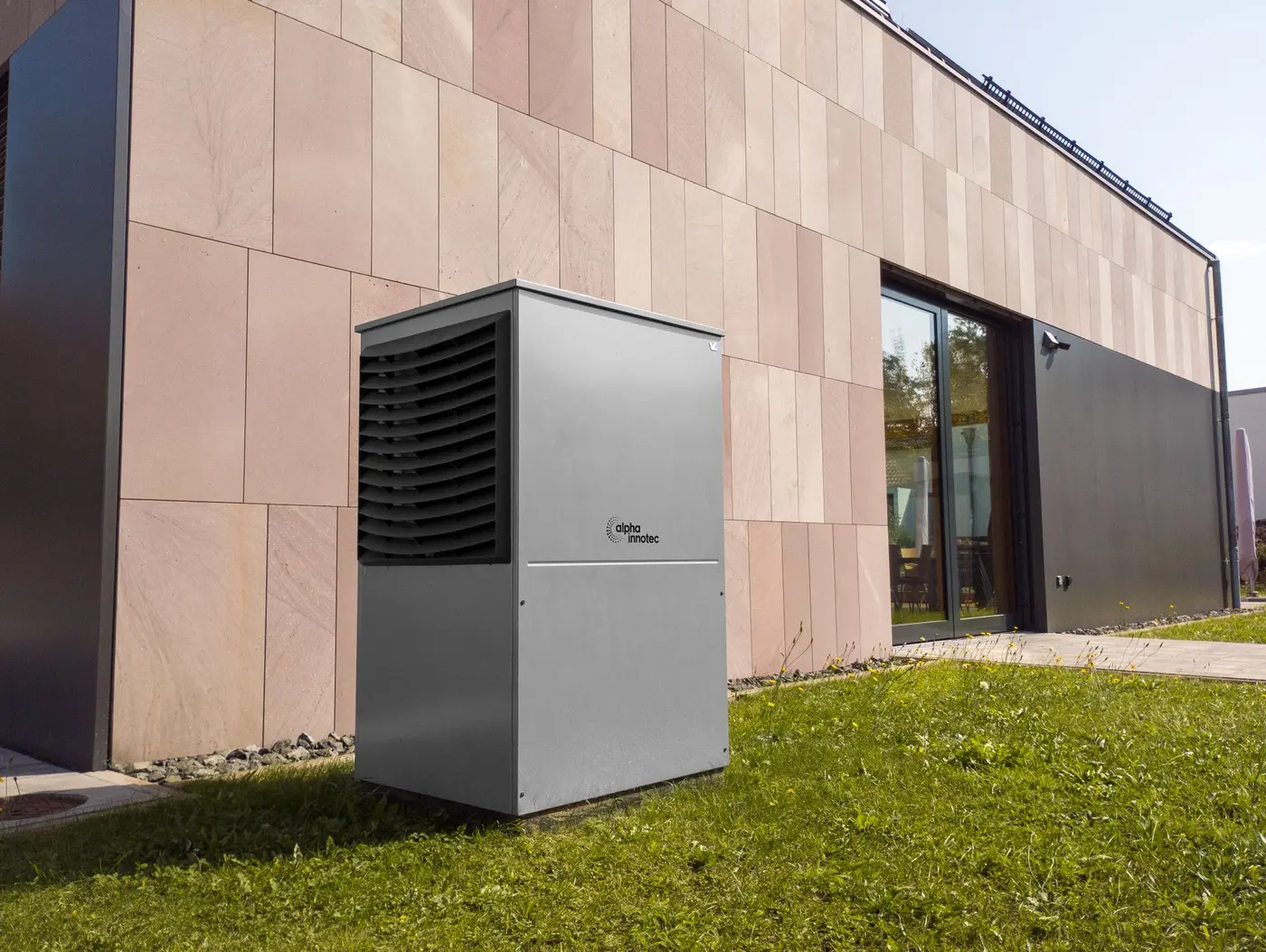 Air-source heat pump condenser standing on a grass strip beside a house wall